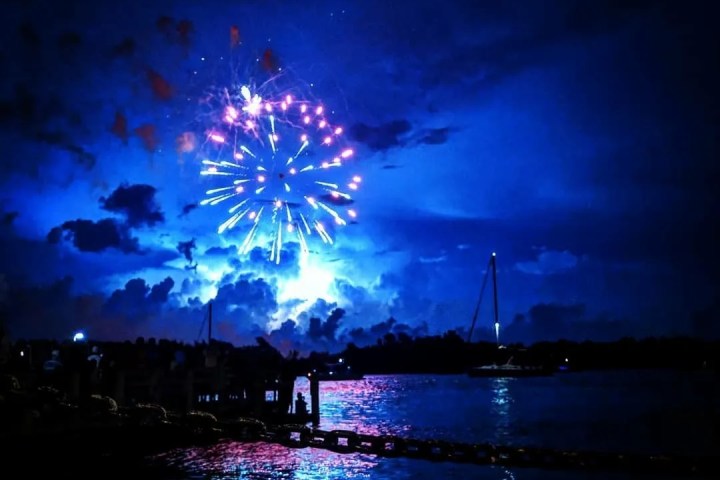 Bright fireworks burst in blue night sky over a calm water surface with reflections.