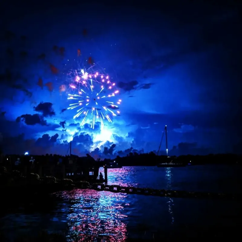 Bright fireworks burst in blue night sky over a calm water surface with reflections.