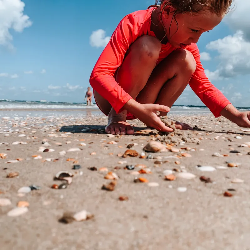 Child in red shirt crouching on a sandy beach collecting shells under a blue sky.